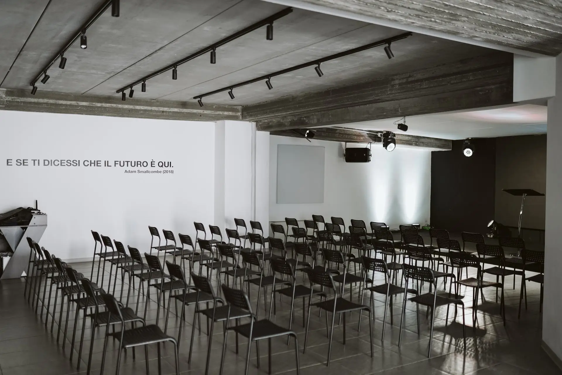 Sala conferenze moderna con file di sedie nere e scritta sul muro bianco: "E SE TI DICESSI CHE IL FUTURO È QUI."