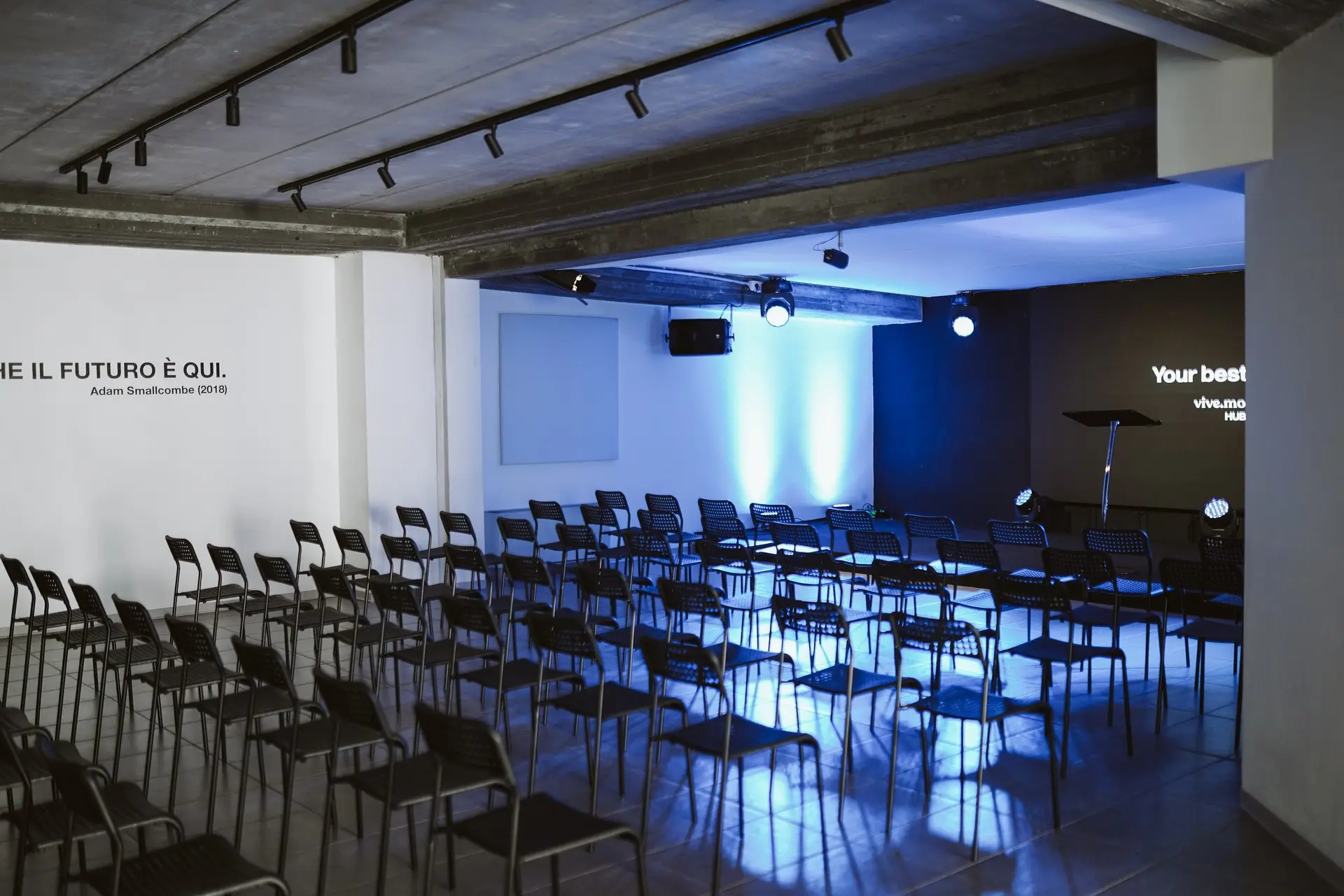 Sala conferenze con file di sedie nere rivolte verso un palco scuro con luci blu