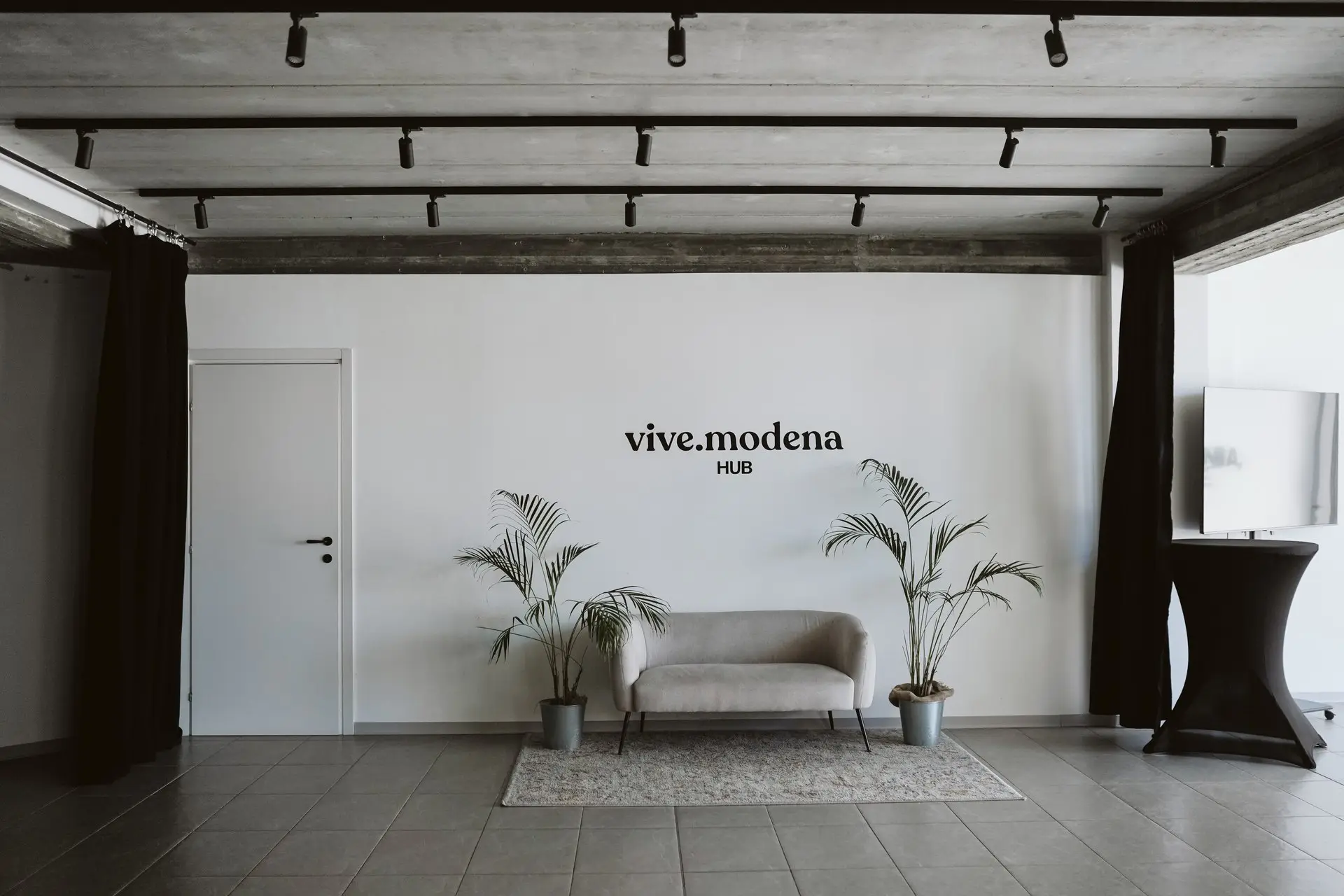 Spazio eventi moderno con logo "vive.modena HUB" su muro bianco, divano grigio e piante