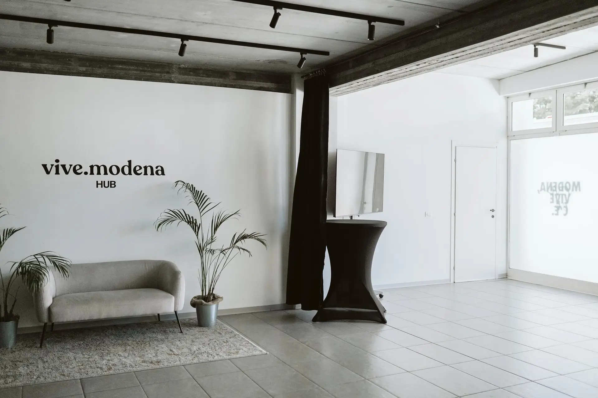 Area reception minimal e moderna con logo "vive.modena HUB", divano grigio e piante