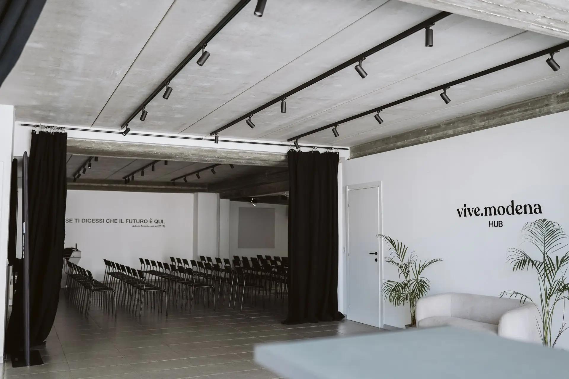Spazio eventi con file di sedie nere, tende nere e logo "vive.modena HUB" sul muro