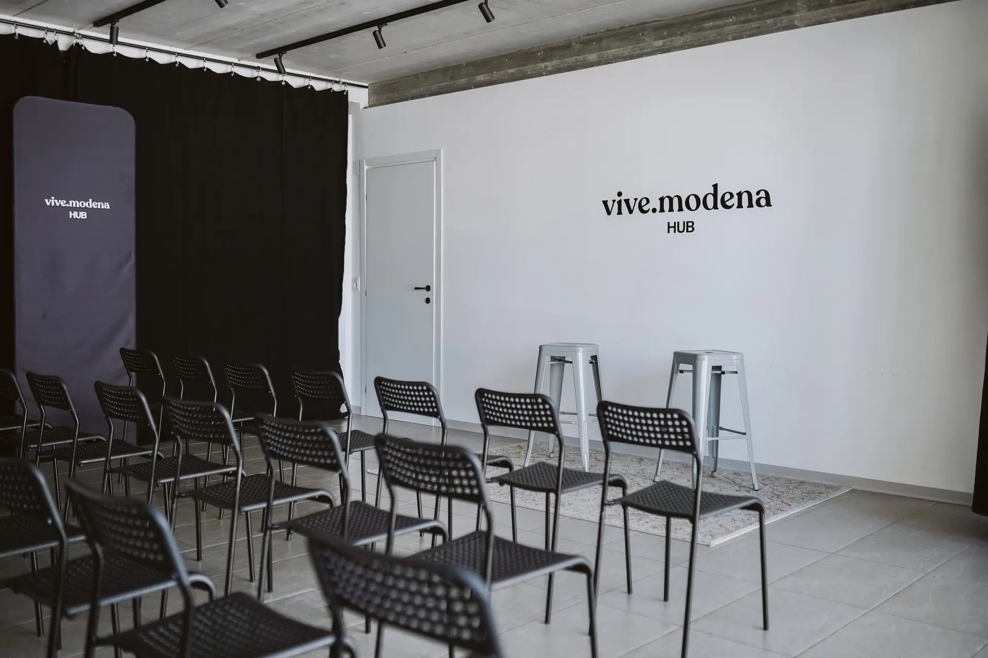 Sala eventi con file di sedie nere rivolte verso un muro bianco con logo "vive.modena HUB"