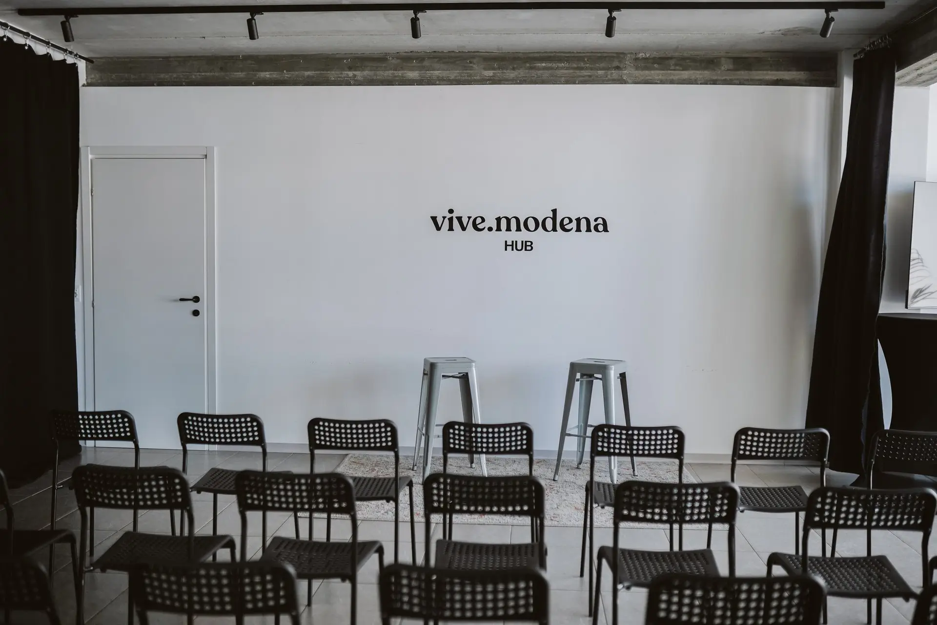 Sala conferenze moderna con logo "vive.modena HUB" sul muro bianco e file di sedie nere