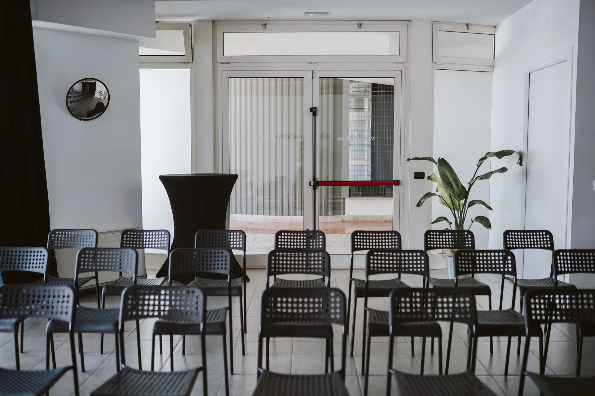 Sala conferenze con file di sedie nere, tavolo alto nero, pianta e porte a vetri